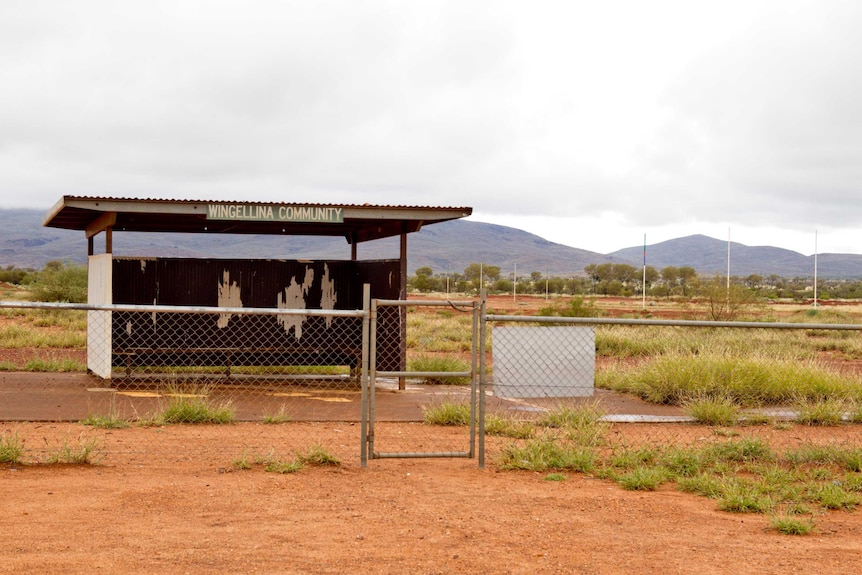 A shelter and a football oval lie behind a wrought iron fence with shrubs scattered on the red dirt.