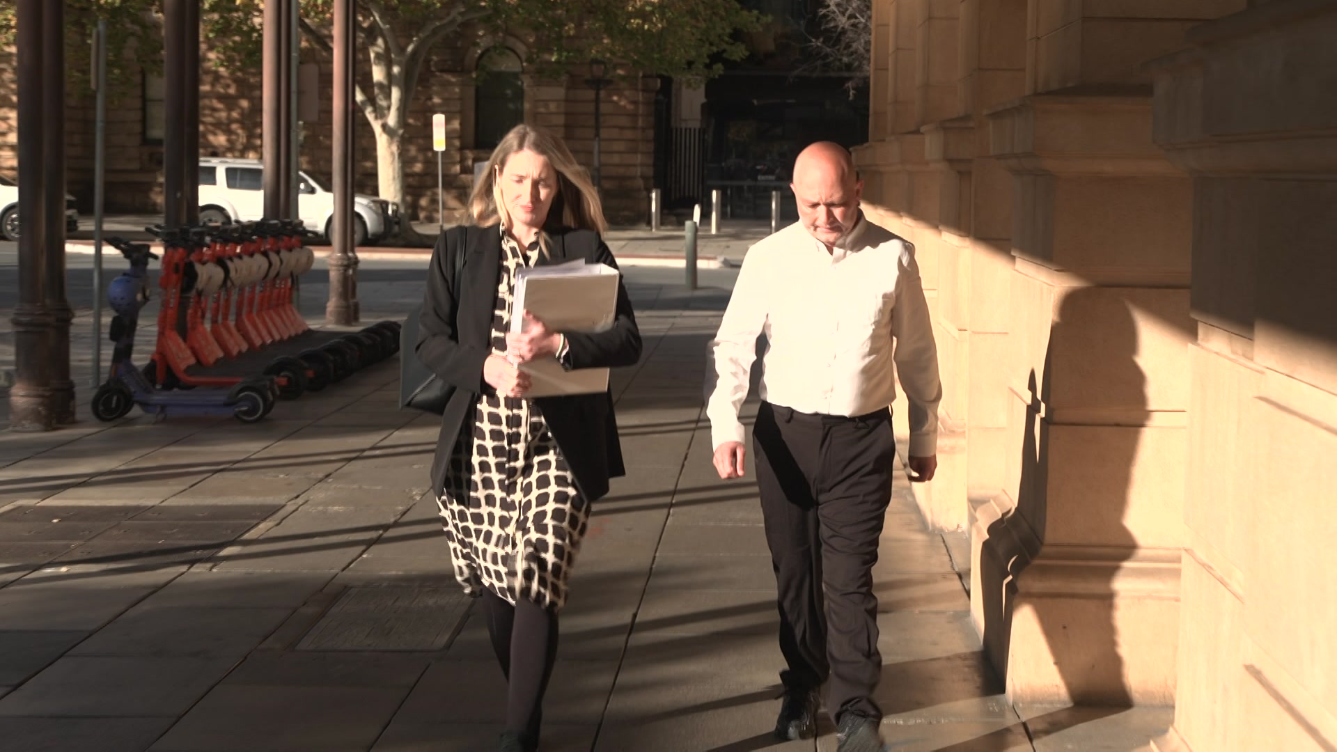 A bald man and a woman wearing a checked dress and black jacket walk on a footpath next to a stone building