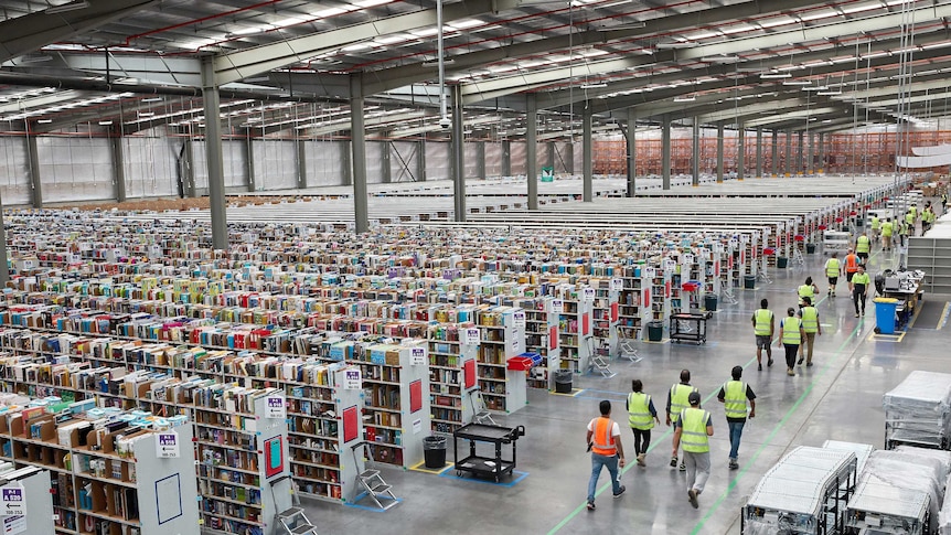 Workers wearing yellow and orange safety vests walk down through the huge warehouse made up of dozens of aisles.