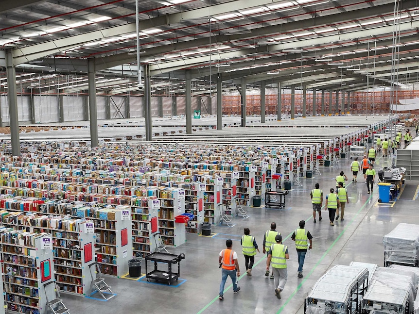 Workers wearing yellow and orange safety vests walk down through the huge warehouse made up of dozens of aisles.