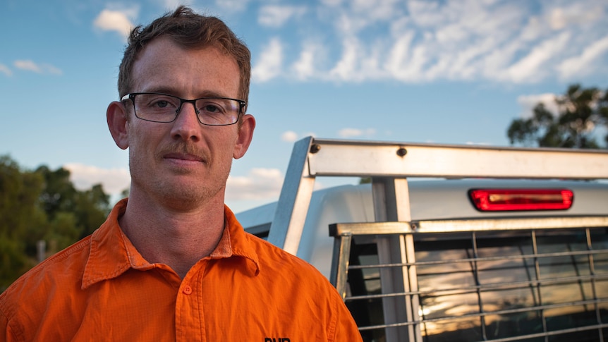 A man wearing an orange shirt stands next to a ute.