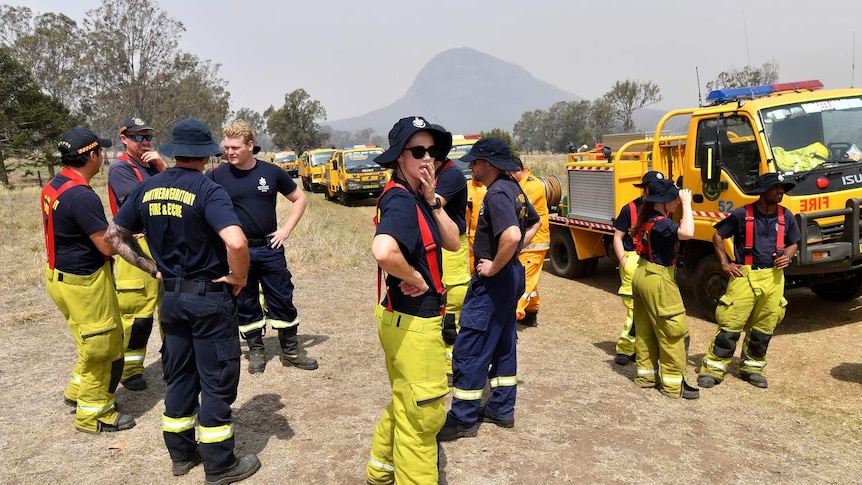 Rural firefighters gather together with hands on hips next to a line of fire trucks, smoky haze surrounds them.