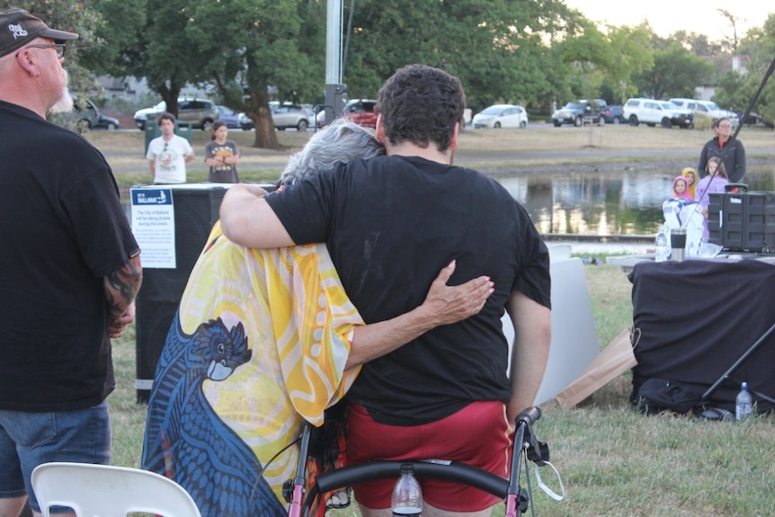 An elderly person and younger person photographed from behind, leaning on each other as they watch a dawn service.