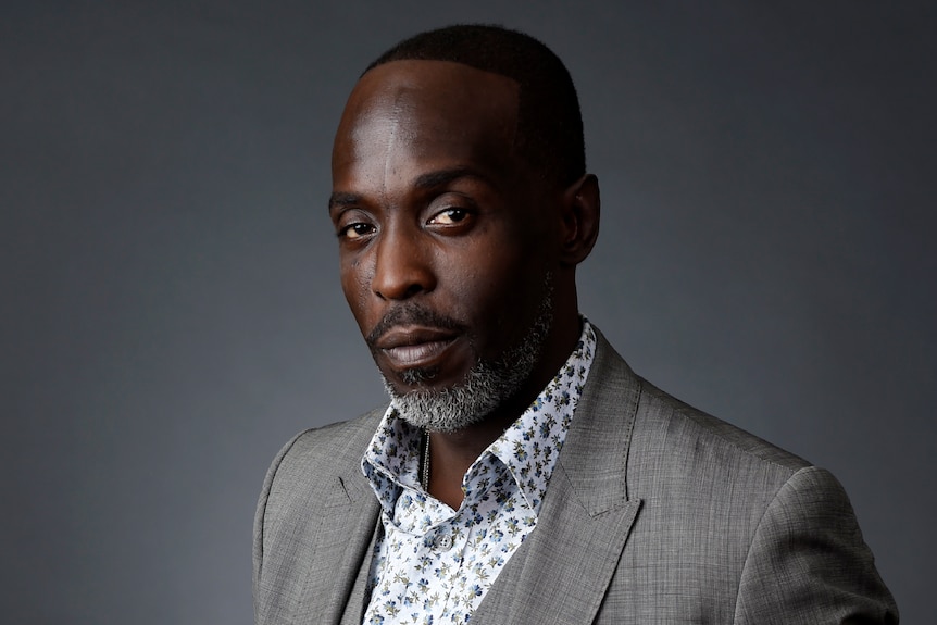 A head shot of actor Michael K Williams, wearing a patterned shirt and grey suit against a grey backdrop. 