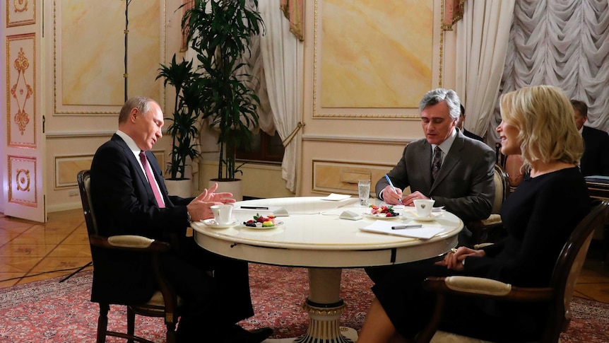 Russian president Vladimir Putin speaks with American brodcaster Megyn Kelly across a table while a translator listens in.