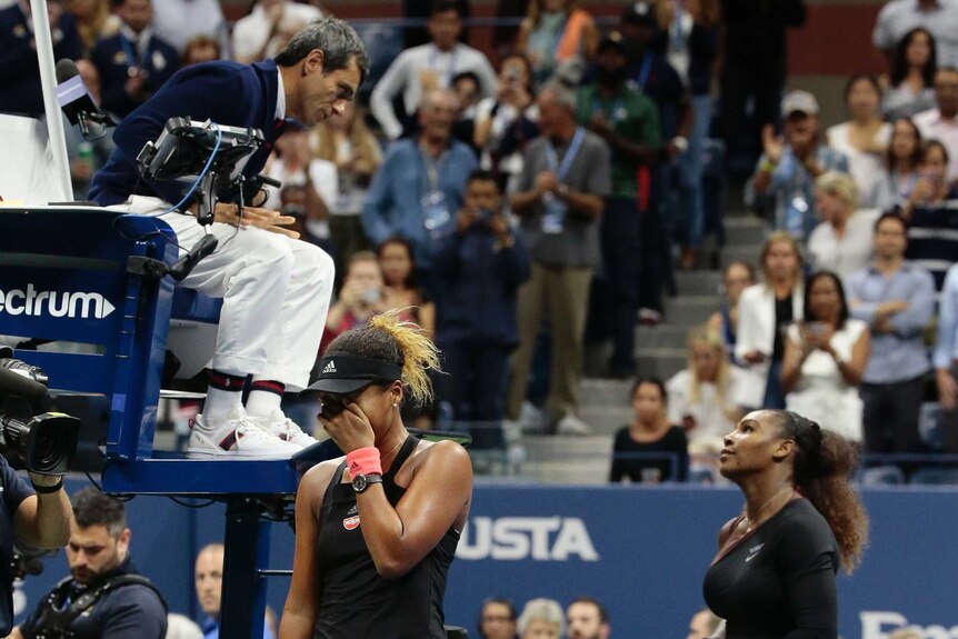 Naomi Osaka walks away covering her face as Serena Williams argues with chair umpire Carlos Ramos.