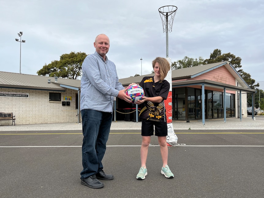 A bald man wearing a blue shirt and jeans with a girl wearing a black shirt and shorts both holding a netball near a hoop