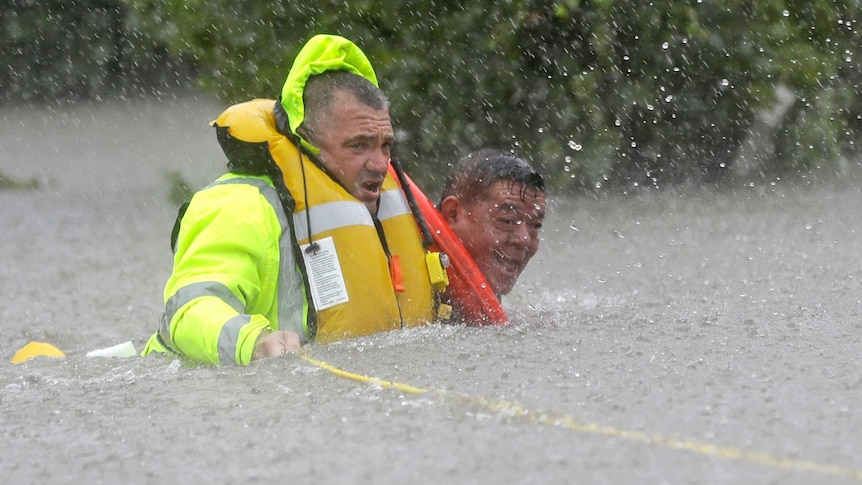 A rescuer in high-vis jacket and inflated life jacket holding onto a rope guides an older man through floodwaters