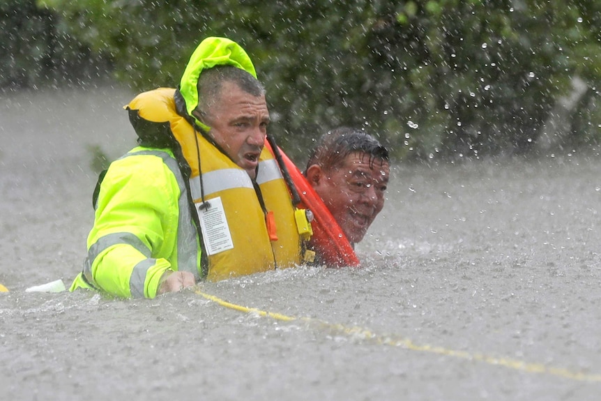 A rescuer in high-vis jacket and inflated life jacket holding onto a rope guides an older man through floodwaters