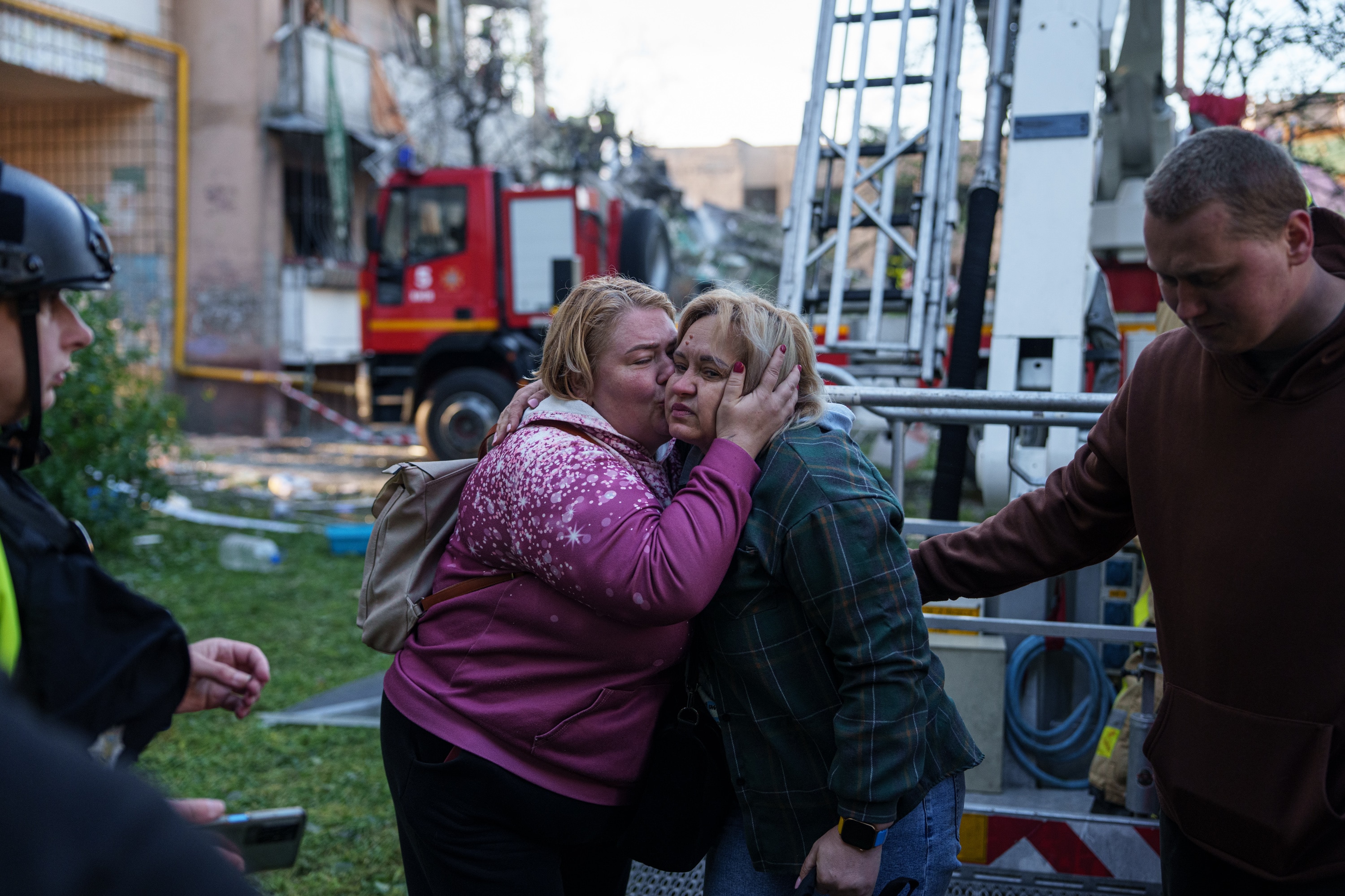 A woman embracing and kissing another woman on the cheek amid rescue workers and firetrucks.