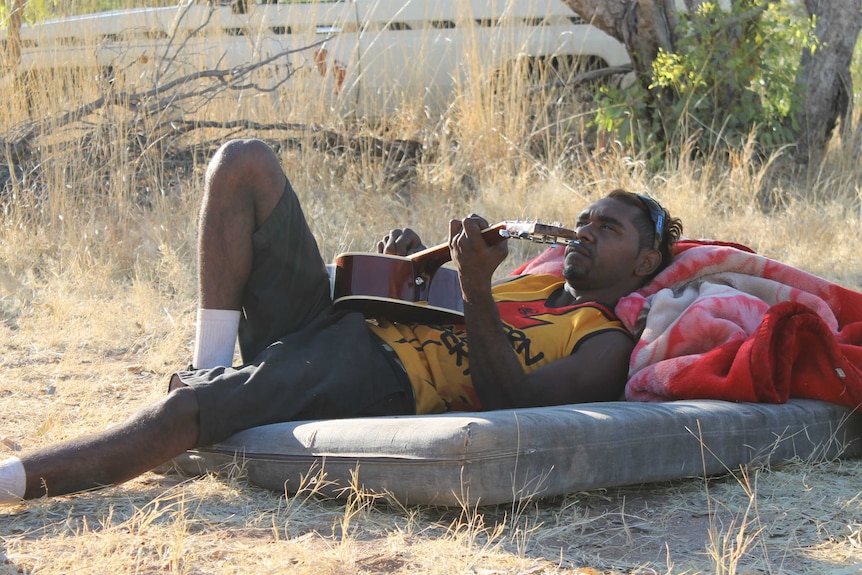 Rocker Wilson playing the guitar while laying outside on a mattress.