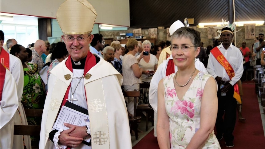 A smiling Archbishop dressed in robes walks down a church aisle with a woman with short, greying hair in white, printed dress.