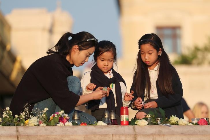 A woman kneels, left, next to two younger girls as they light candles among flower tributes.