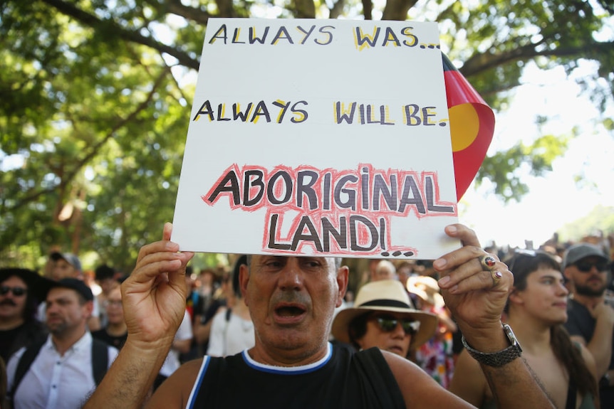 A man holds a sign that says 'Always was.. Always will be Aboriginal Land.'