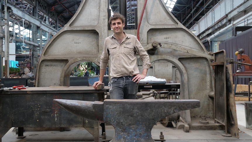 A man standing behind an anvil in a industrial workshop.