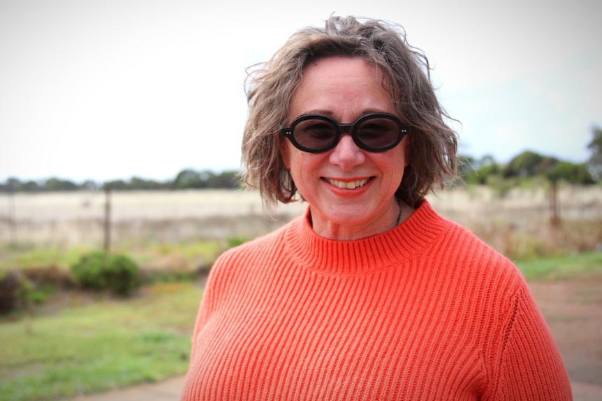 A woman in an orange jumper stands in front of a paddock. She has curly hair and black glasses.