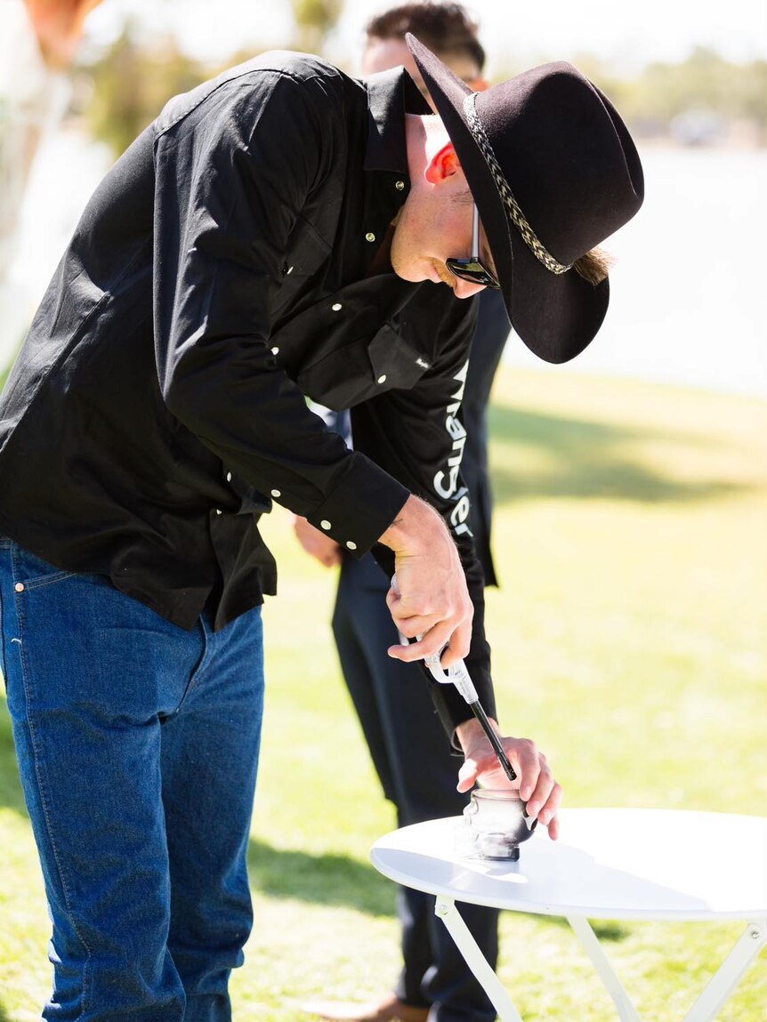 A man in a hat, sunglasses and a black shirt lights a candle on a sunny day