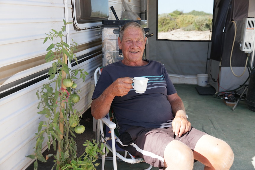 An older man sits outside his caravan, holding a cup of tea and smiling.