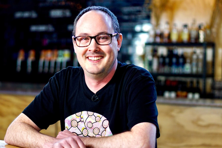 A man in a black t-shirt with glasses smile at the camera, as he leans on a counter with beer bottles behind him.