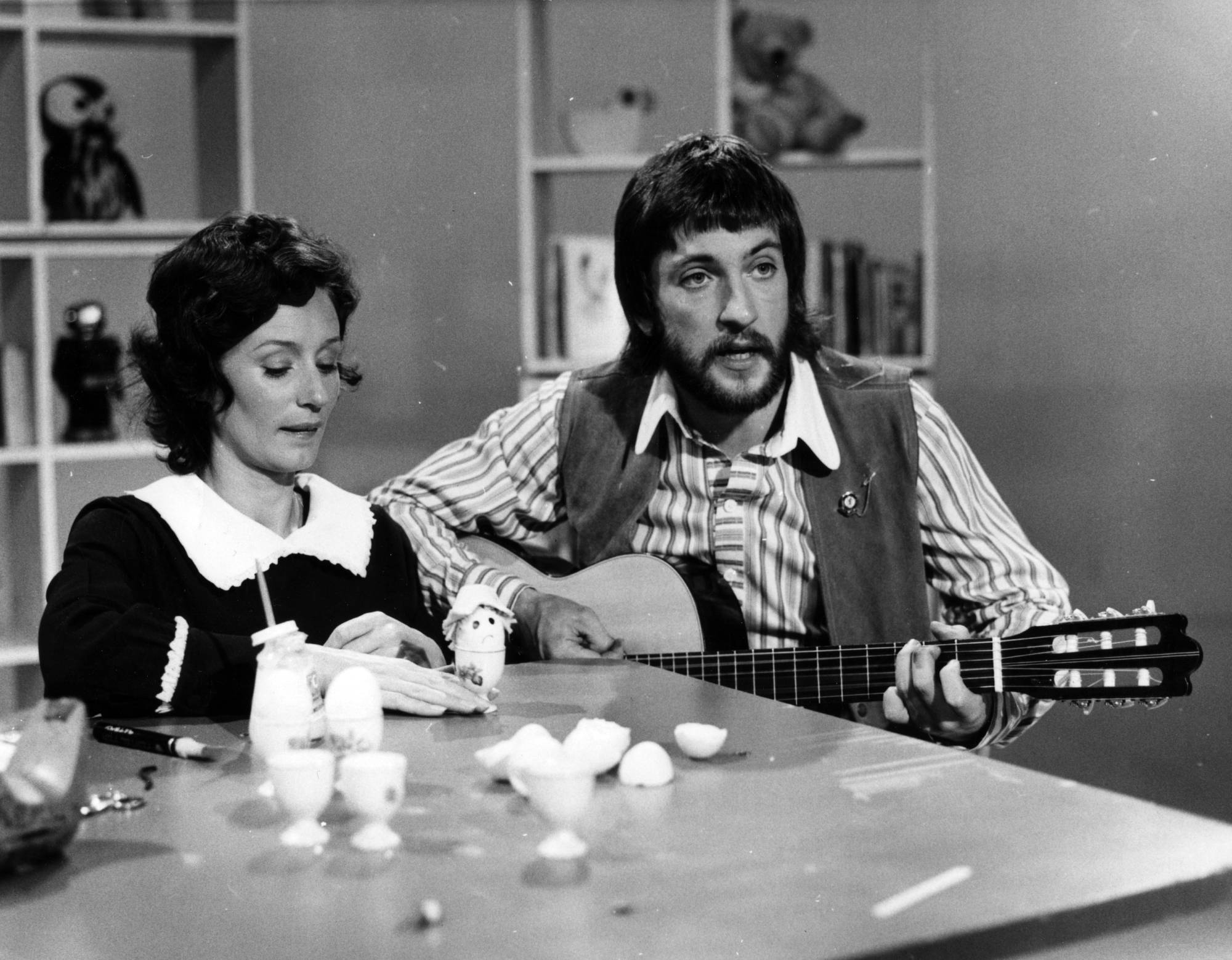 A black and white photo of Benita Collings and John Waters.  John is playing a guitar and Benita is playing with a toy.