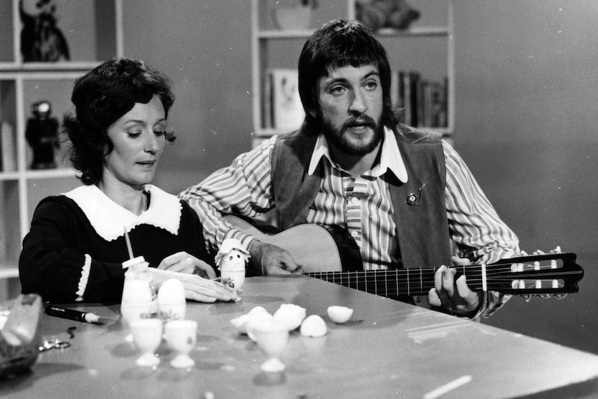 A black and white photo of Benita Collings and John Waters. John is playing a guitar and Benita is playing with a toy.