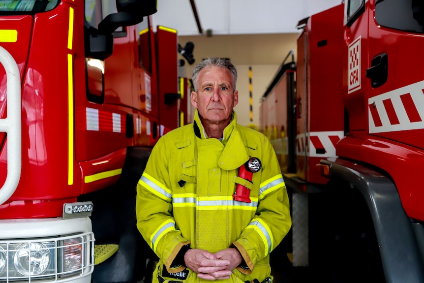 A man in bright yellow fire gear stands between two red fire trucks. 