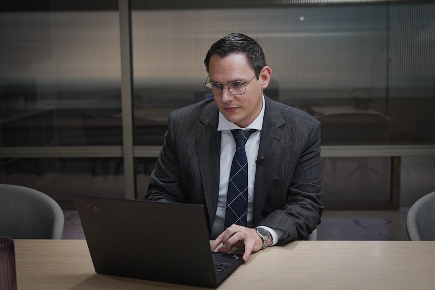 a man with short hair and glasses in a business suit and tie types on  laptop in a corporate office