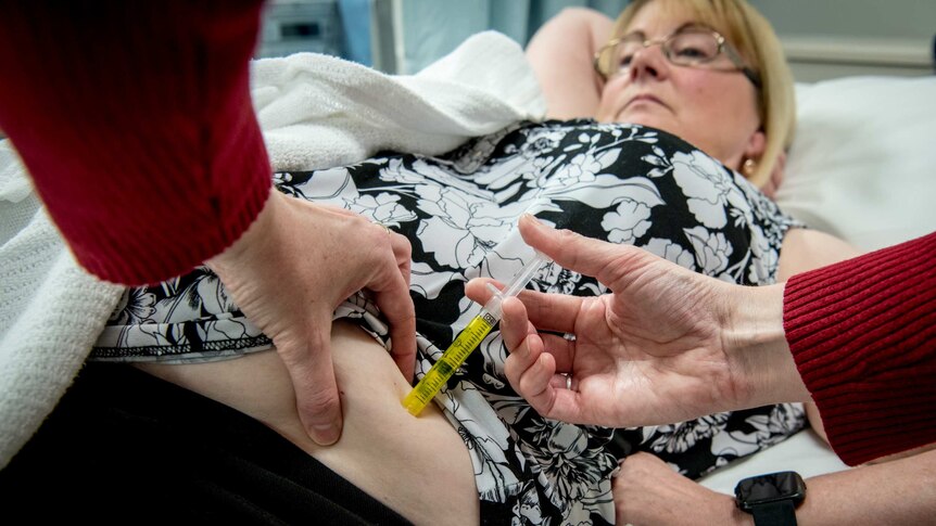 Karen Cooke being injected with the trail a drug whilst laying down on the bed.