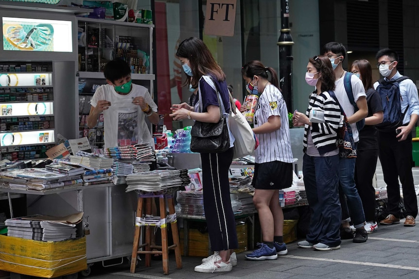 People line up at a newspaper stall in a city street.