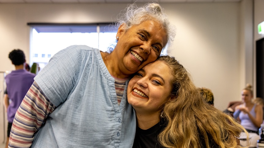 A young Indigenous woman with blonde air and black shirt embraces an elderly Indigenous woman with white hair and blue jacket.