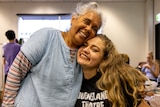 A young Indigenous woman with blonde air and black shirt embraces an elderly Indigenous woman with white hair and blue jacket.