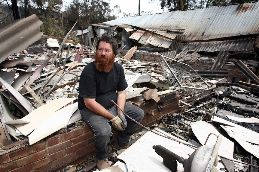 A man sits among the charred remains of his house in the wake of a bushfire.