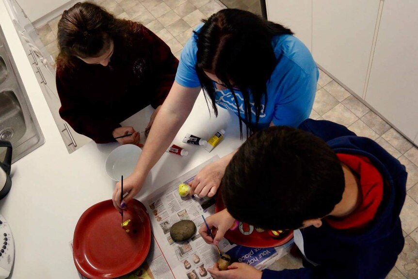 Three children in care in Canberra decorate rocks in a kitchen.