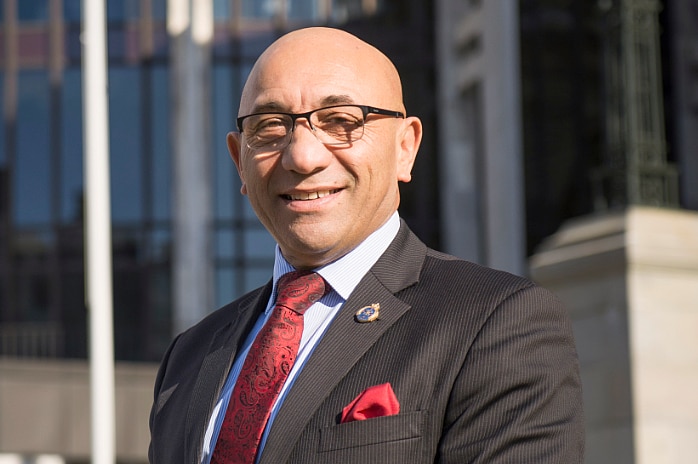 A bald man in a black suit and red tie with tanned skin smiles at the camera.