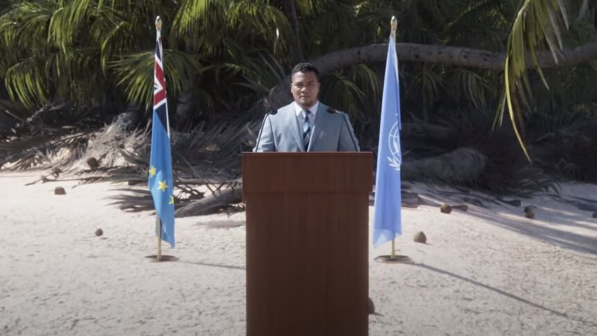 Man in front of podium with flags and beach sand behind him.