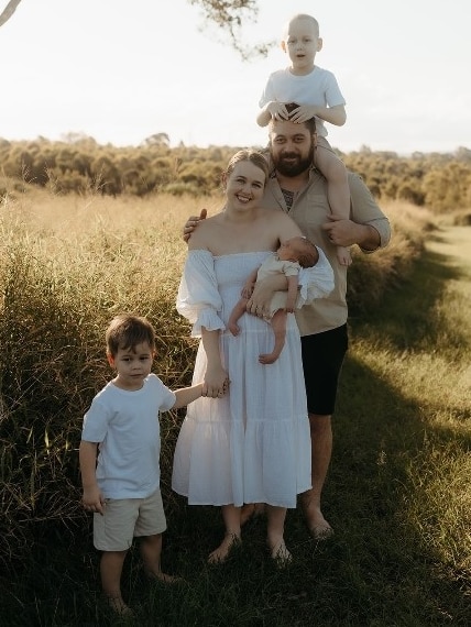 A family of three children and two adults pose smiling together in a field with a sunset in the background.