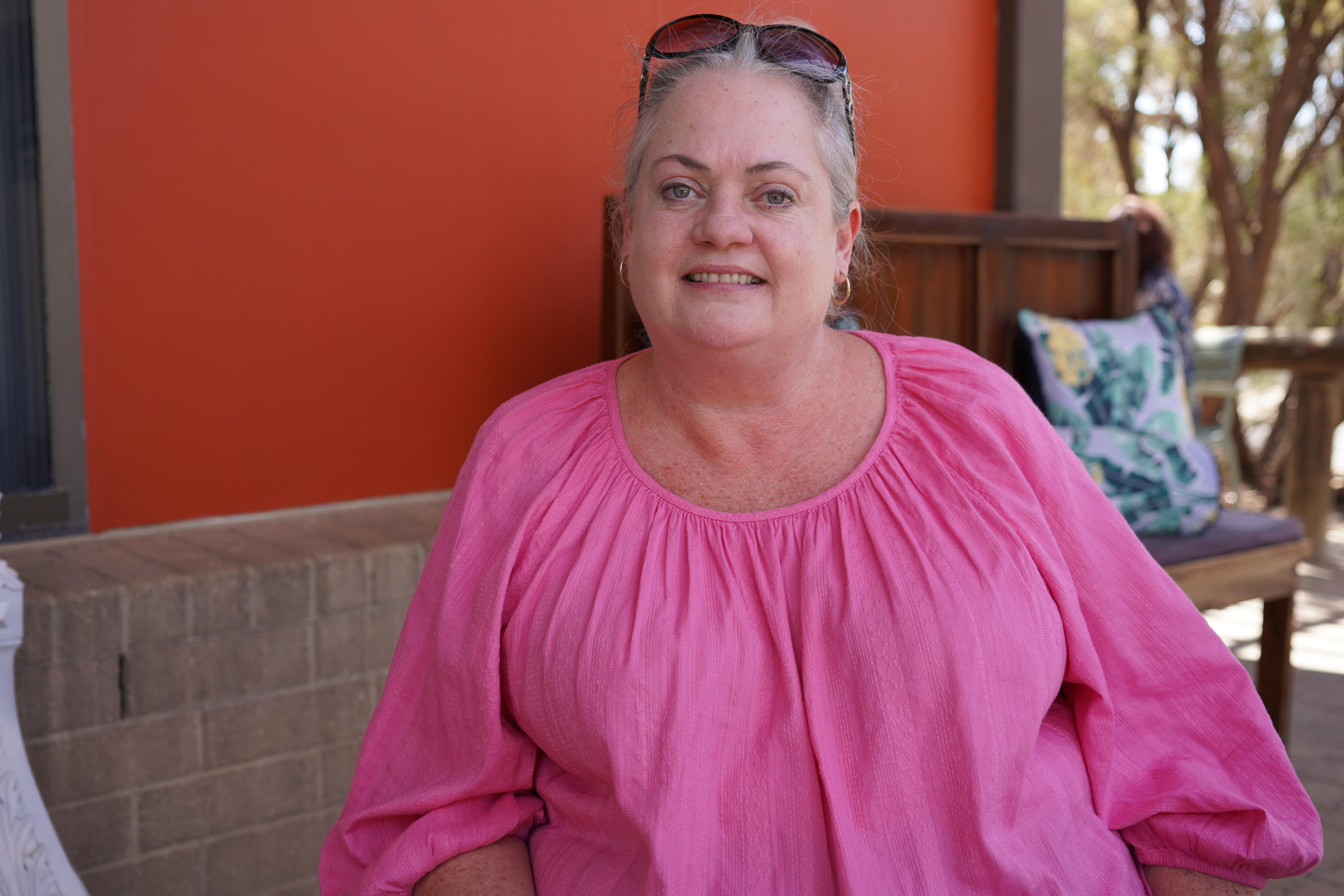 A woman in a pink top sits outside at a cafe smiling at the camera. 