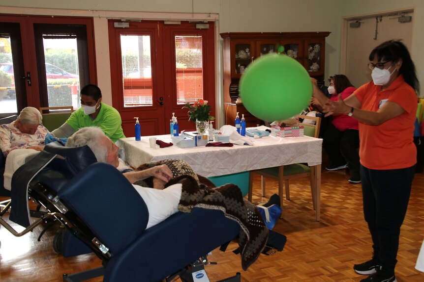 A carer with a green balloon and aged care home residents in chairs