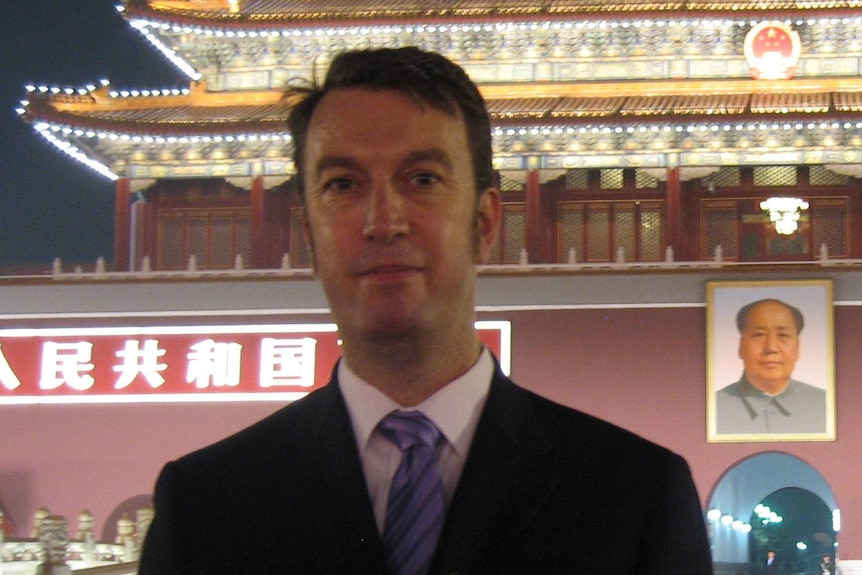 A headshot of a man in a suit in front of an elaborate temple lit up with a big picture of Mao Zedong.