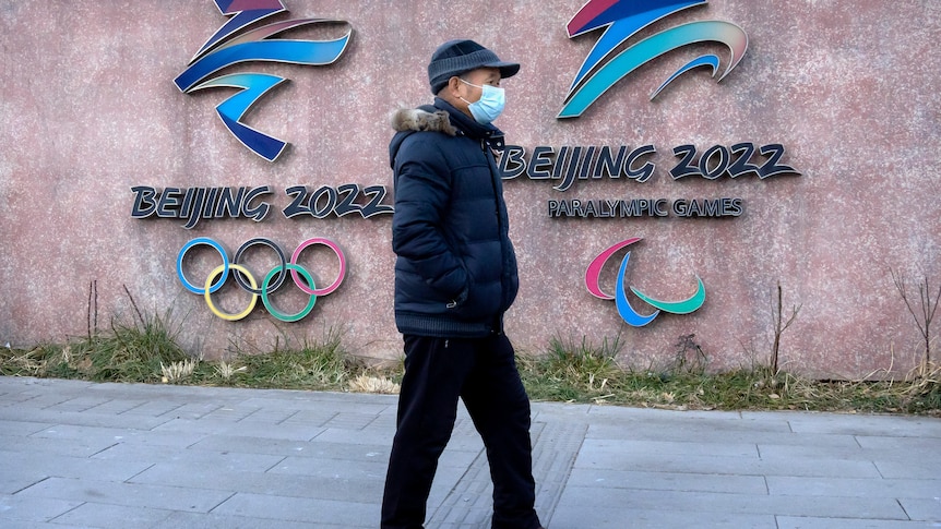 A man in a mask walks past two signs with Beijing 2022 logos featuring the Olympic rings