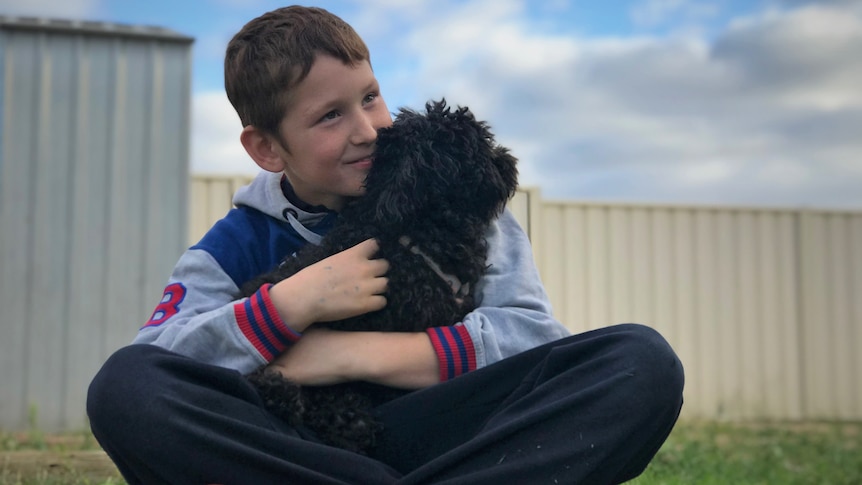 A young boy holding a dog and smiling into the distance. There's a blue sky behind him.