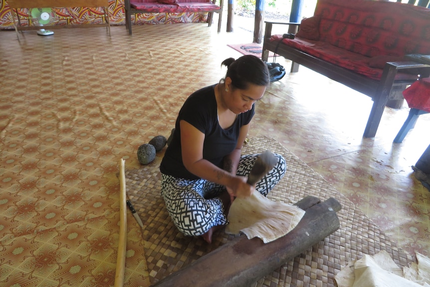 A woman sits on a bark cloth mat using a brush to smooth a piece of wood