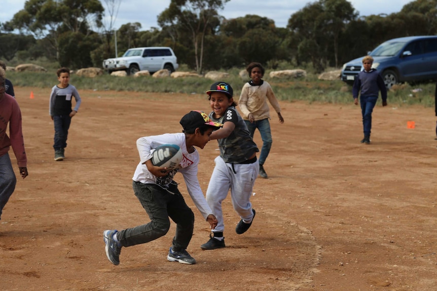 Young aboriginal kids play with a footy ball on a red dirt oval.