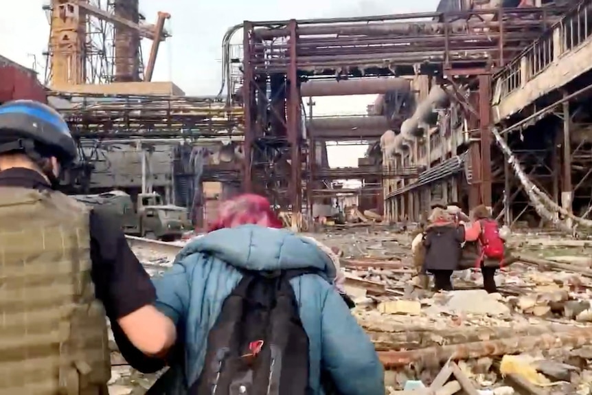 A soldier helps a person walk by supporting her arm while others walk ahead over rubble and under industrial infrastructure