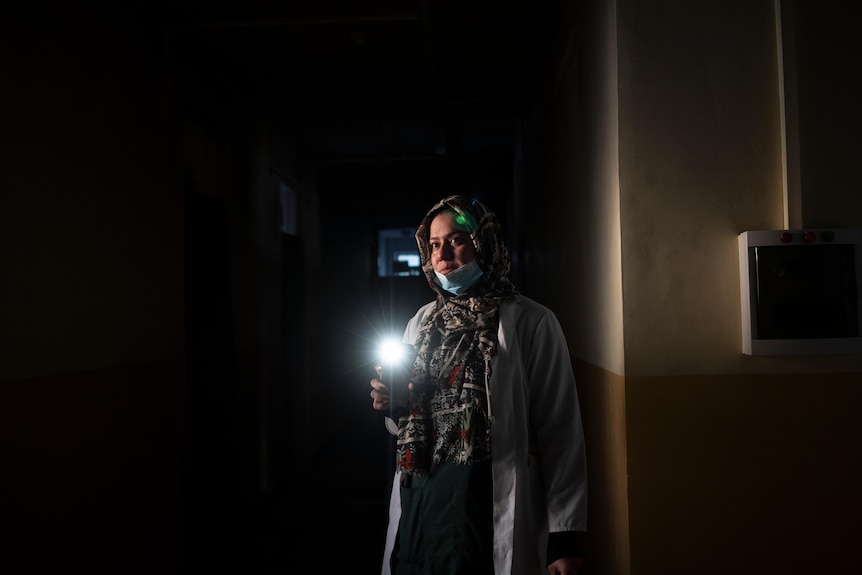 A Middle Eastern woman in head scarf shines a torch in a darkened corridor inside a hospital