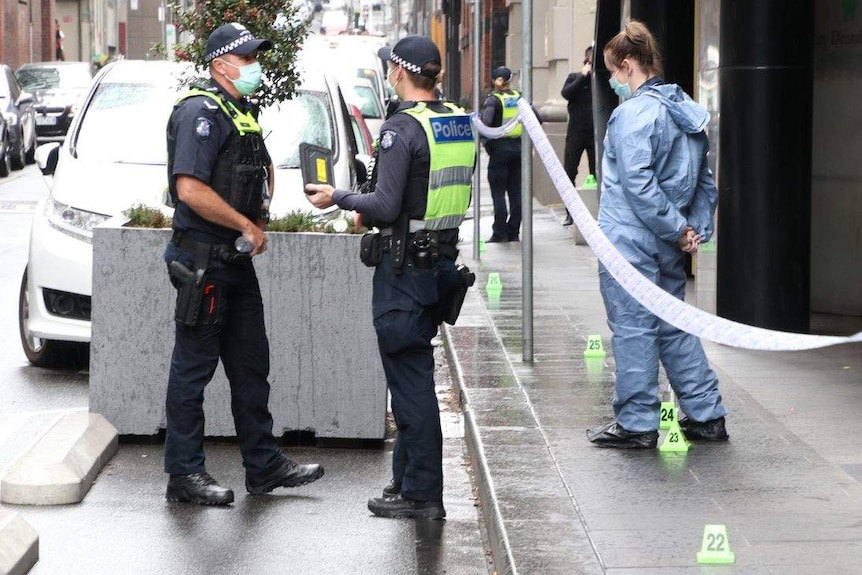 Two police officers and an investigator standing on a footpath with police tape and green crime scene evidence markers.