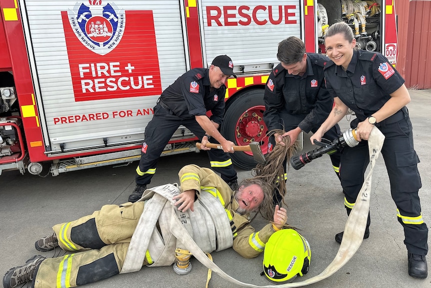 A Fire and Rescue NSW crew pose with Jason Baldock wrapped in a fire hose, pretending to chop his hair with an axe.