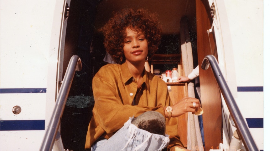 A film still of a young Whitney Houston, sitting on the outside steps of a private plane.
