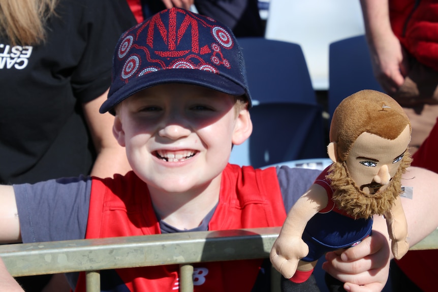 A young Demons fan smiles while holding a Max Gawn doll at Melbourne training before the AFL grand final.