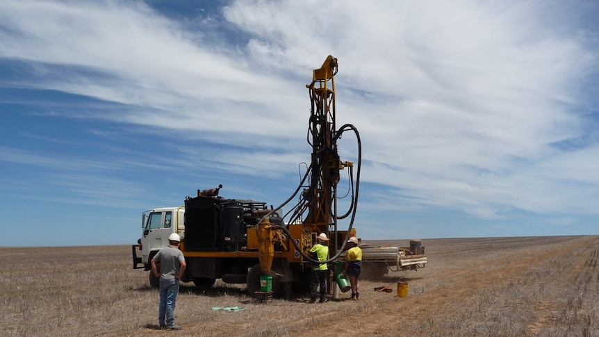 A drilling rig mounted on the back of a small truck sits in an empty field, three men are operating it.